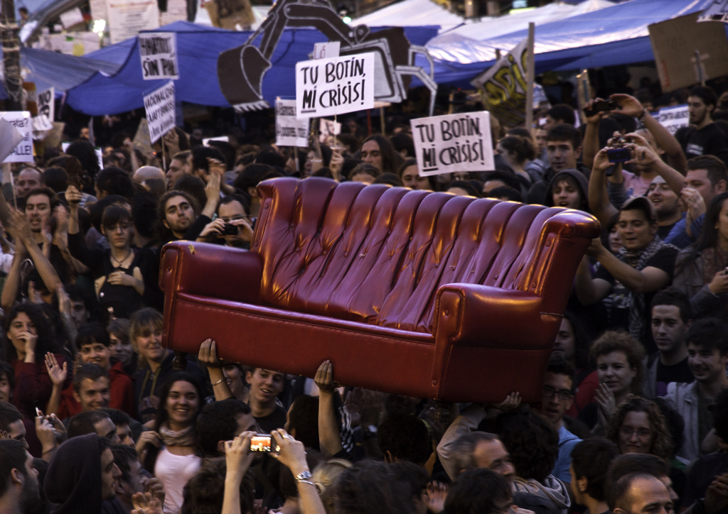 Trabajar gratis. Plano general, color. Manifestantes del 15M en Plaza Sol (Madrid) llevando en volandas un sofá rojo.
