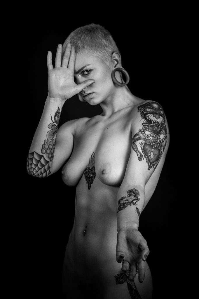 Plano americano, blanco y negro. Modelo, desnuda y tatuada, tapandose media cara con la mano y mostrando el tatuaje de su antebrazo. 