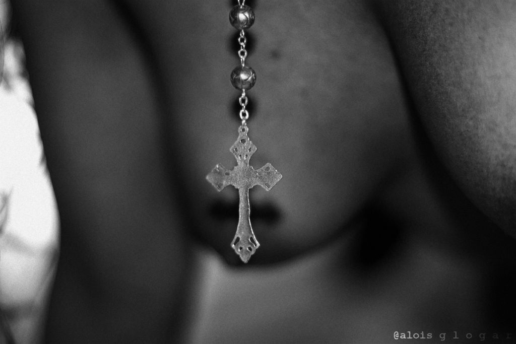 Escandalizarse es de ignorantes. Plano medio, blanco y negro. Crucifijo de rosario en primer plano. De fondo pecho mujer desnuda desenfocado. 
