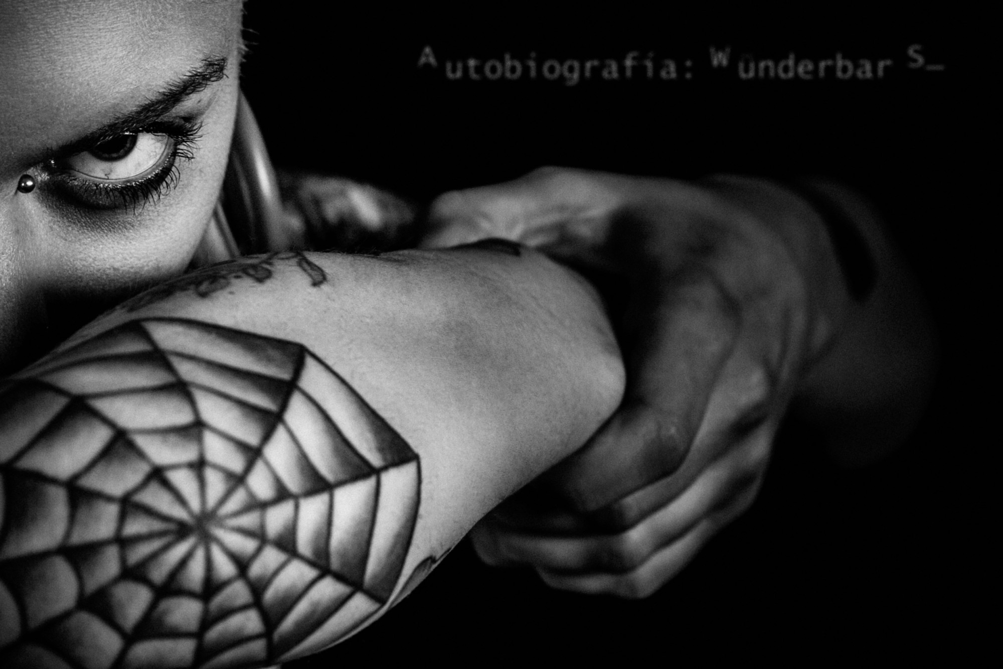 Primer plano, blanco y negro. Modelo con tatuaje de tela de araña en el codo, poniendose el brazo como un antifaz.