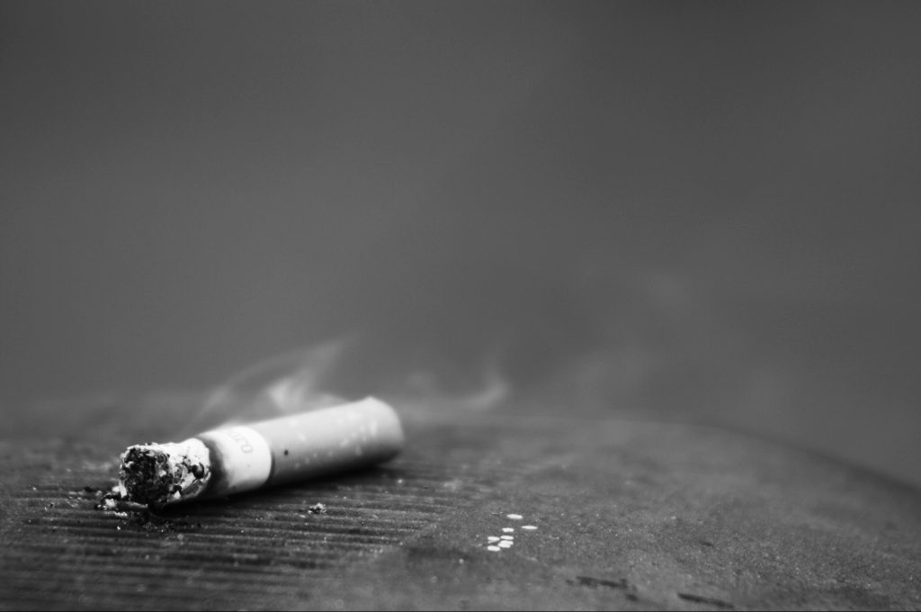 Resumen del 2016. Plano general, blanco y negro. Cigarrillo abandonado sobre una papelera callejera consumiéndose con un hilo de humo.