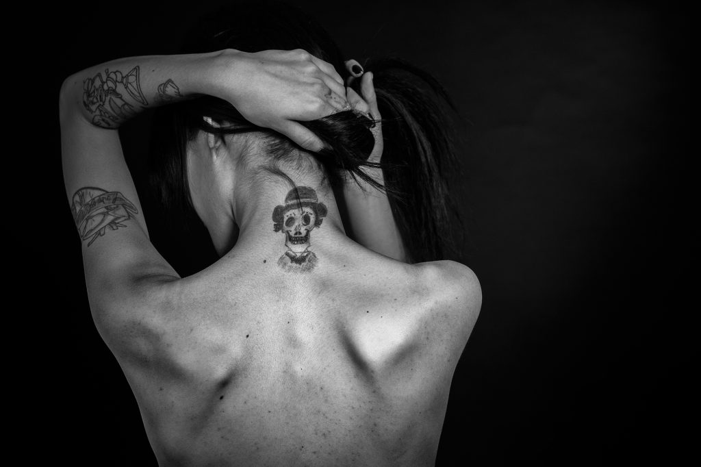 Fotografía y realidad. Mujer desnuda, en blanco y negro. De espaldas, sujetándose el pelo para mostrar un tatuaje en su nuca. 