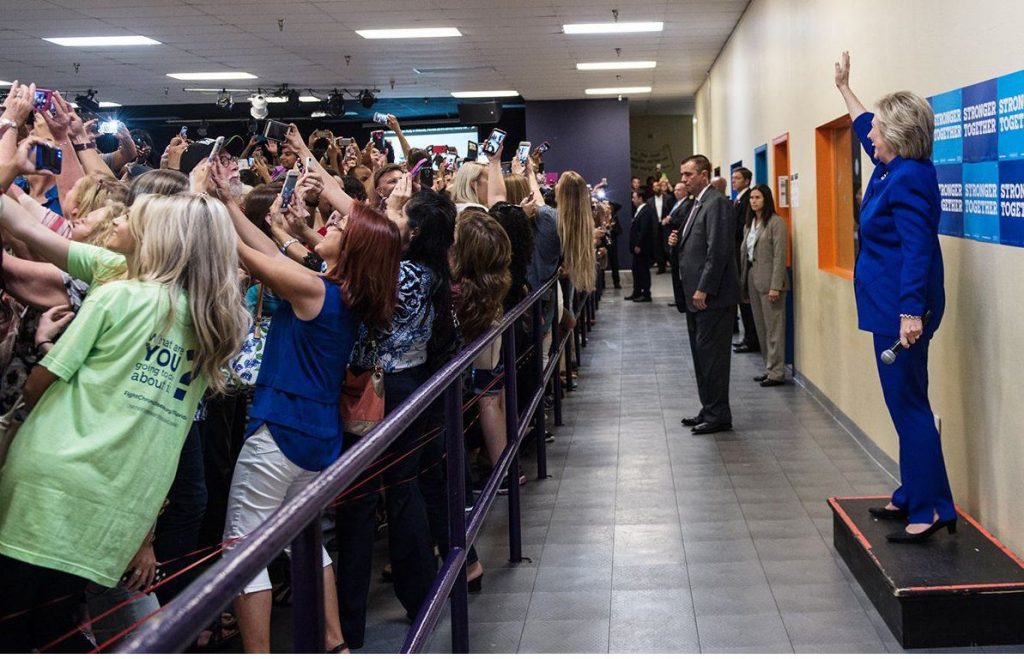 Mirada fotográfica. Plano general, color. Hillary Clinton pidiendo a su público que se haga selfies con ella de fondo. 