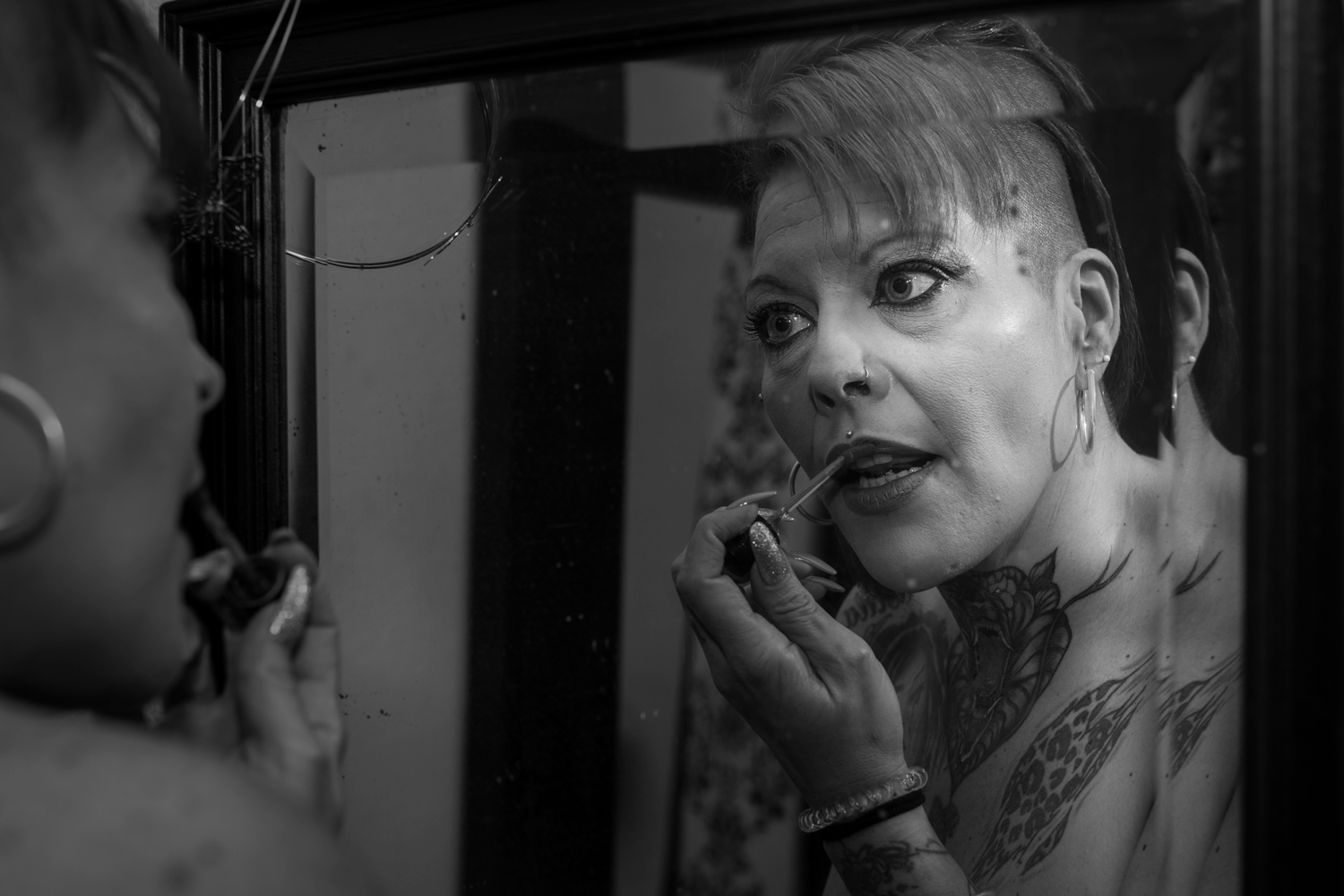 Primer plano, blanco y negro. Modelo tatuada y vieja, maquillándose delante de un espejo roto.