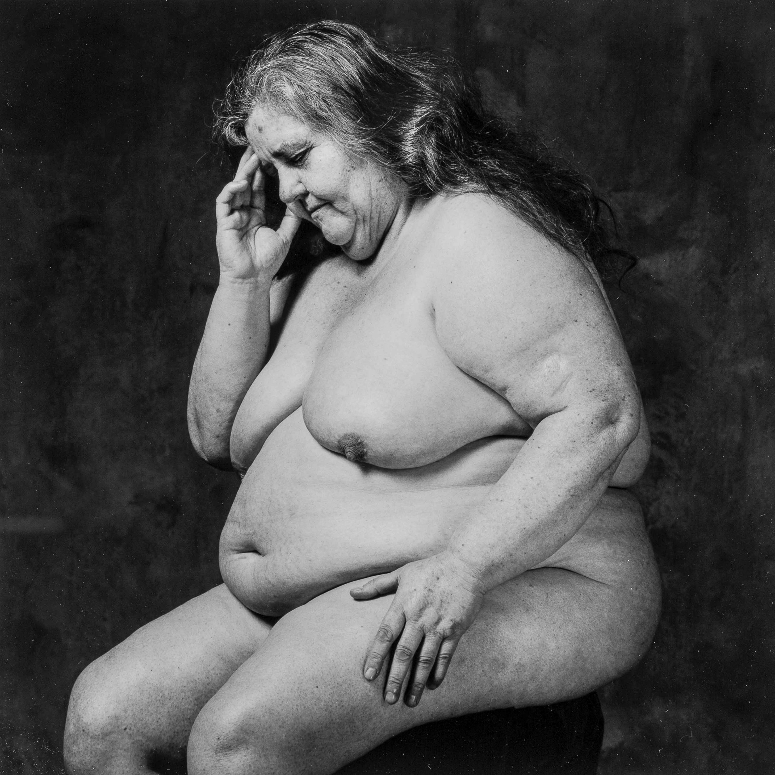 Plano americano, blanco y negro. Mujer desnuda, con sobrepeso mórbido, sentada en actitud que recuerda al pensador de Rodin.