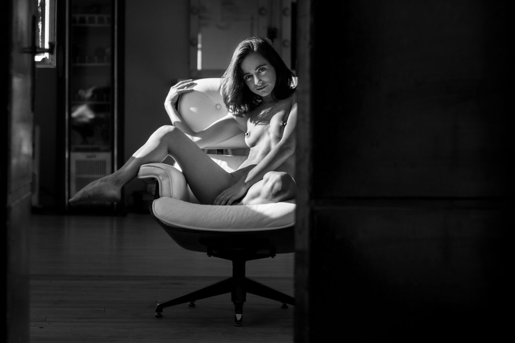 Plano general, blanco y negro. Chica desnuda sentada en un sillón con actitud sugerente. vista a traves de una puerta entreabierta. 