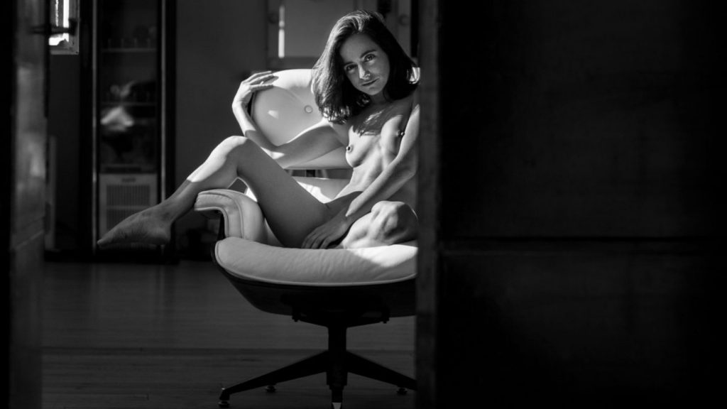 Lo más visto en la web en 2020. Imagen en blanco y negro. Mujer desnuda sentada en un sillón. Vista a través de una puerta entreabierta. Mira sonriendo al espectador