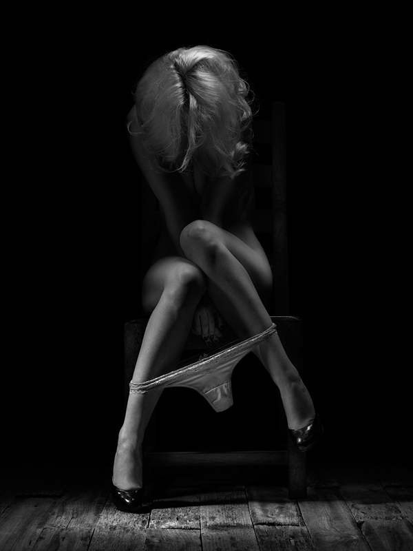 Imagen en blanco y negro. Mujer desnuda, sentada y emergiendo de un fondo totalmente negro. Lleva zapatos de tacón y bragas a la altura de los tobillos. 